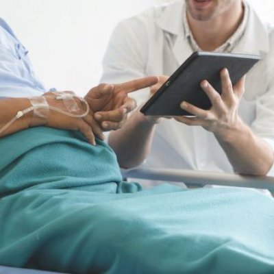 A patient sits up in a hospital bed and points at a tablet being held by a clinician sitting beside them.